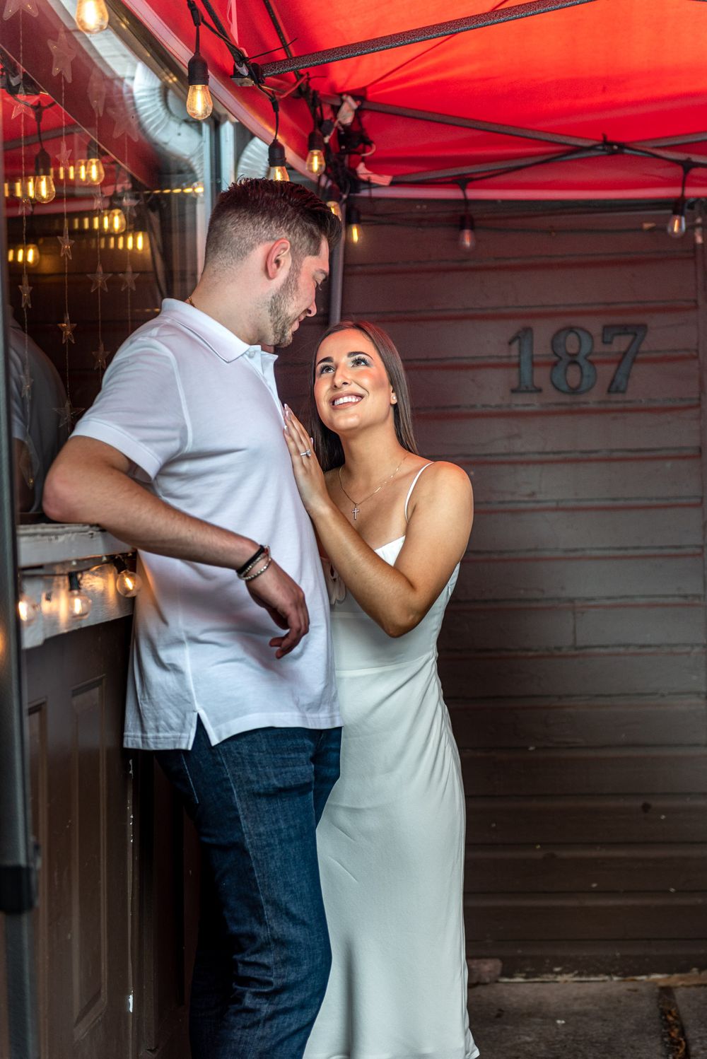 Couple sharing a romantic moment under string lights at address 187, woman in white slip dress smiling up at man.