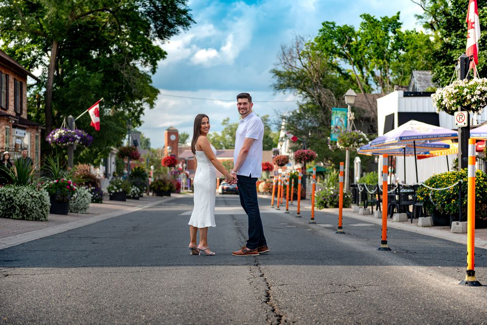 Couple holding hands while posing on a charming flower-lined street with Canadian flags in the background.