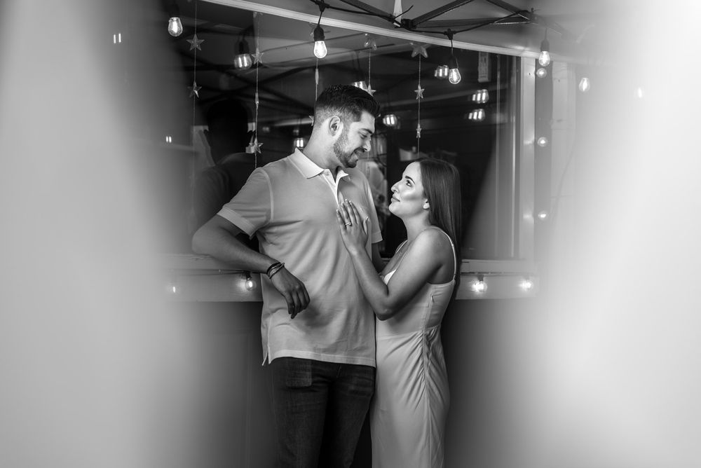 Black and white photo of couple gazing at each other under hanging lights in a romantic urban setting.