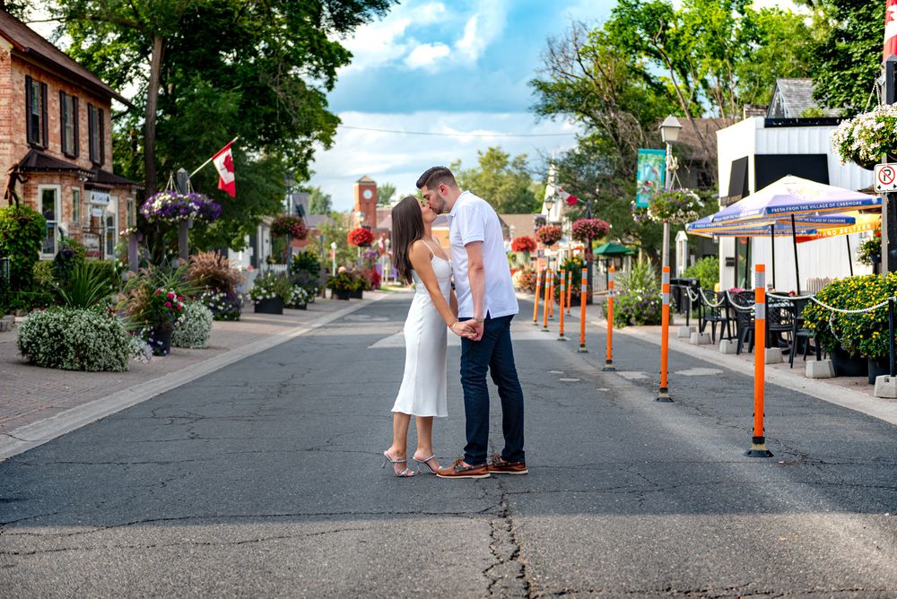 Couple embracing on a picturesque flower-decorated street under a bright blue sky with fluffy clouds.