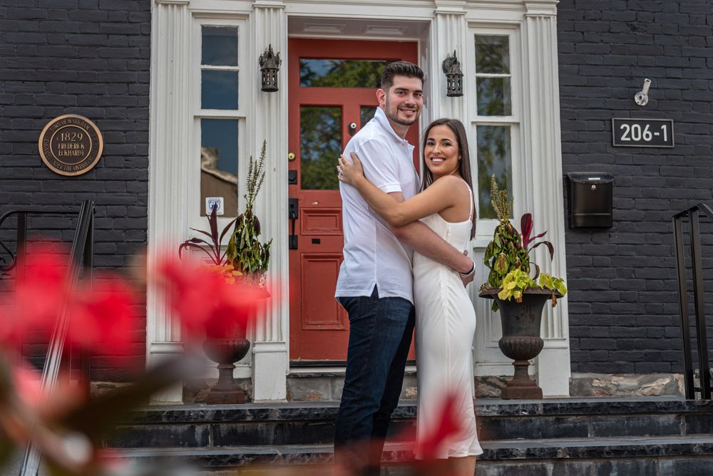Happy couple hugging on steps of an elegant black house with red door, surrounded by blooming red flowers.