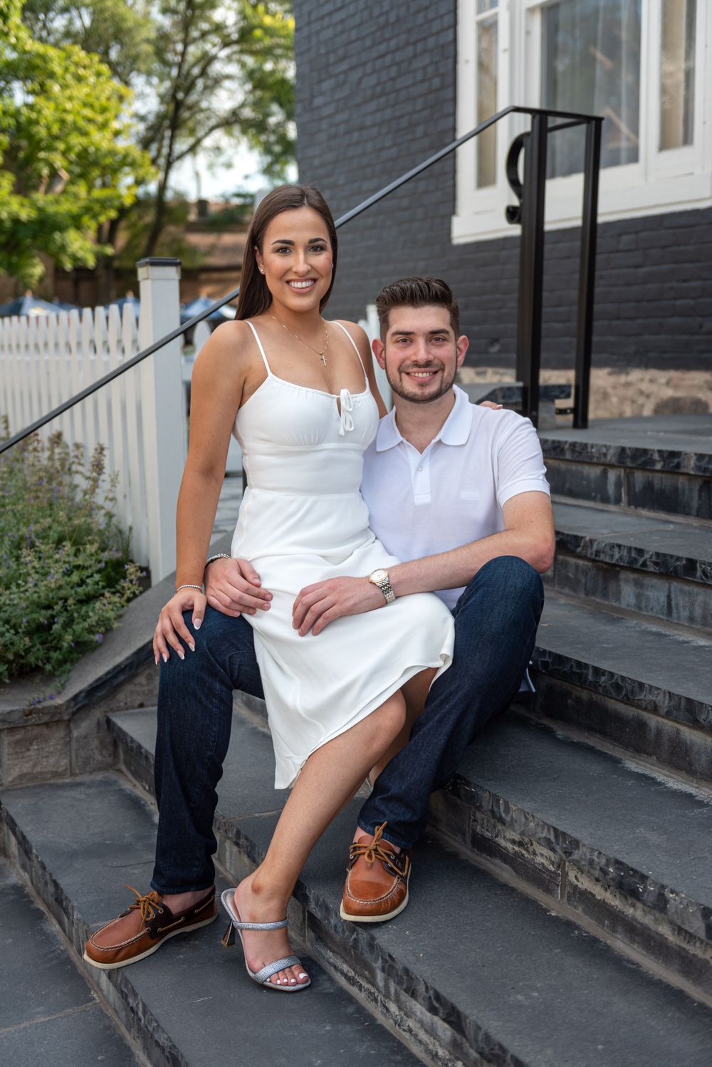 Smiling couple in white outfits sitting on stone steps outside a dark brick building for an engagement photo.