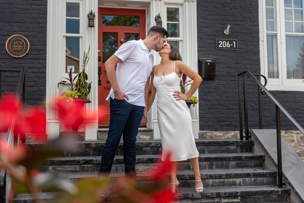Couple sharing a romantic kiss on steps of a dark house framed by red flowers in the foreground.