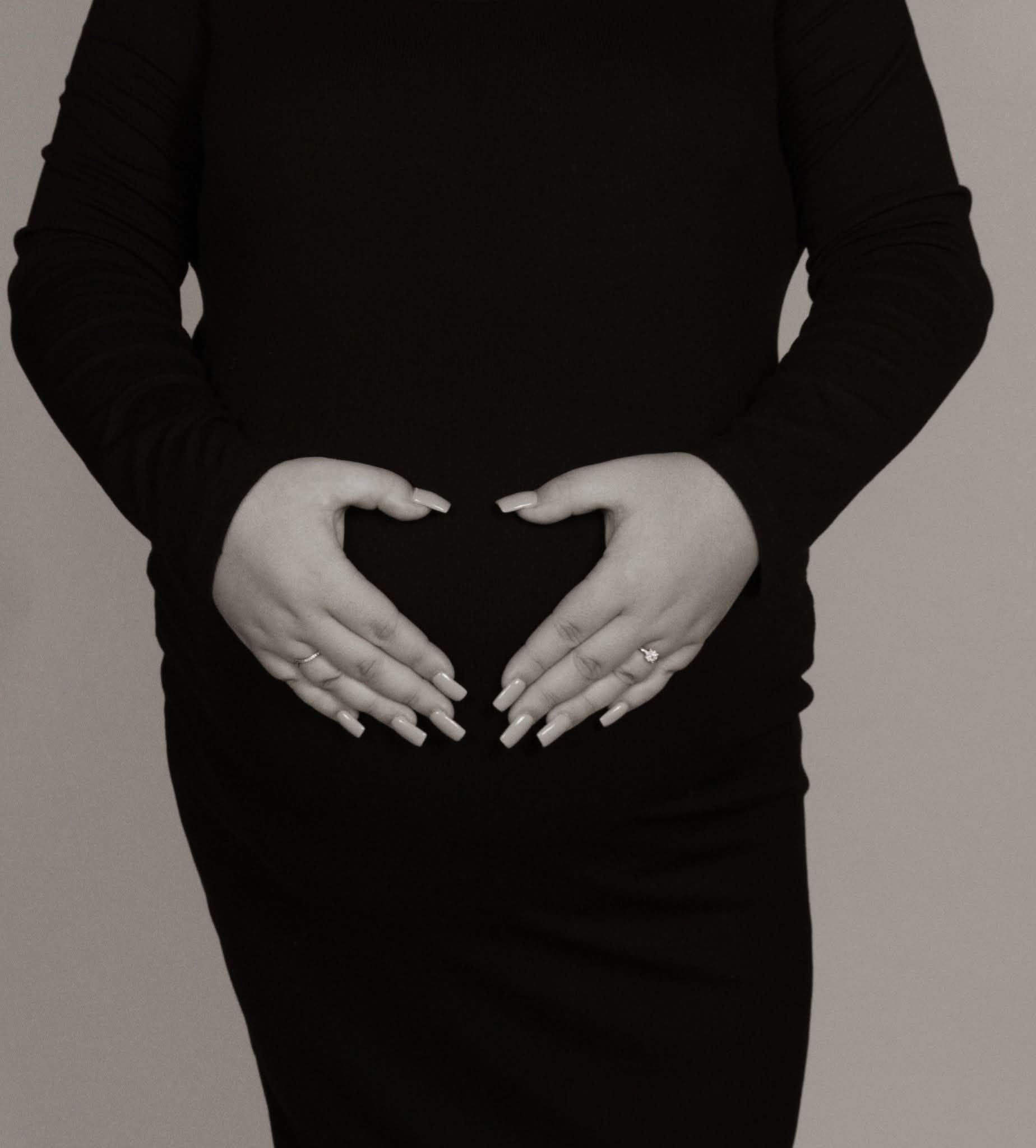 Black and white maternity photo of pregnant woman in black dress forming heart shape.