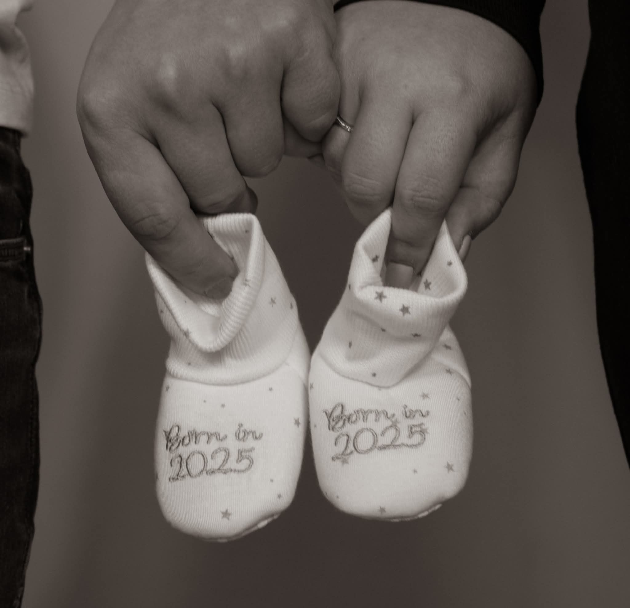 Two hands holding tiny baby booties embroidered with Born in 2025 in black and white.