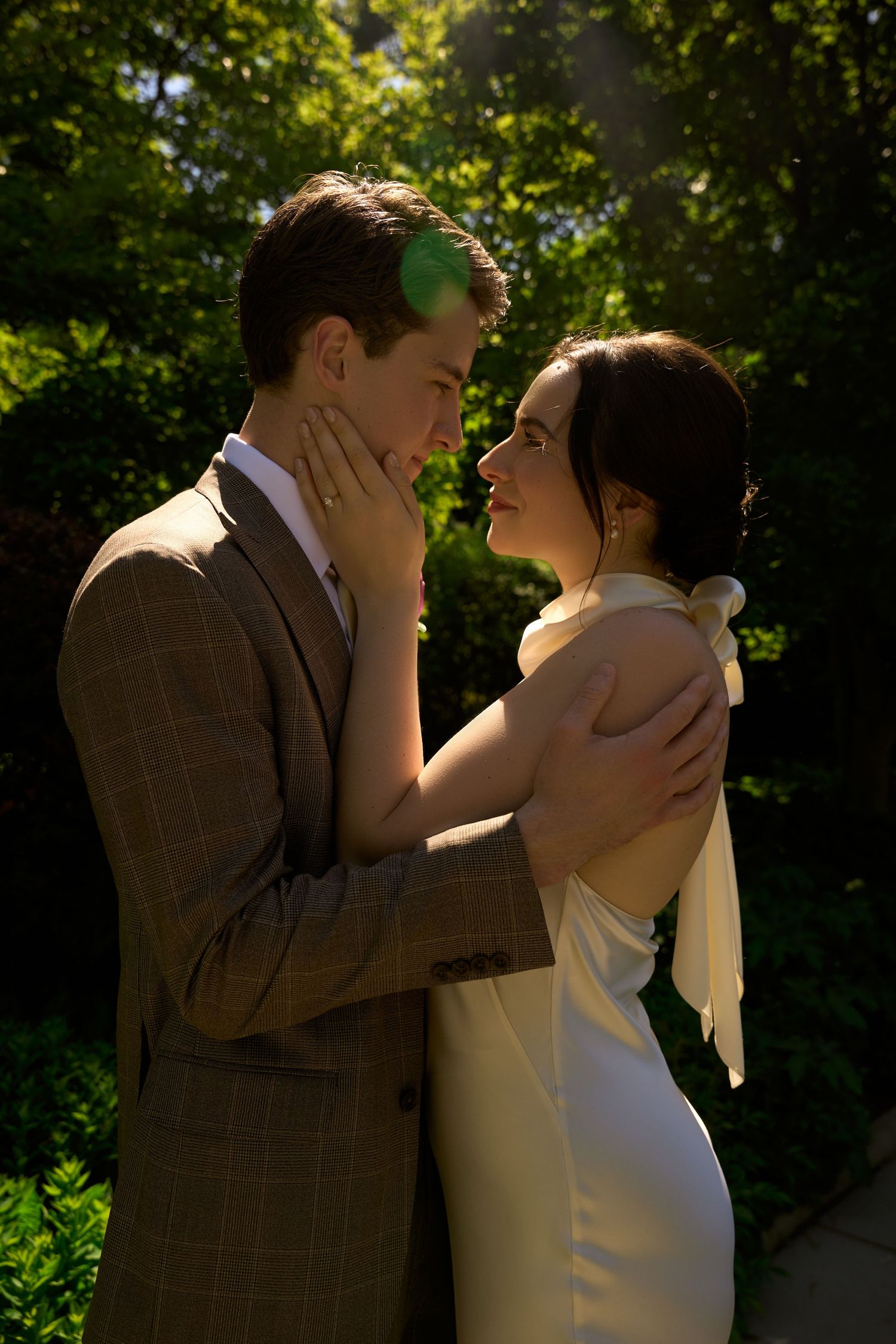 Couple in romantic embrace outdoors, groom in brown suit, bride in white dress, surrounded by lush green foliage in soft sunlight.