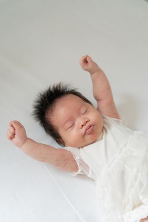Peaceful newborn baby sleeping in white outfit with arms raised, dark hair, on white background.