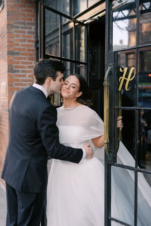 Bride and groom share a tender kiss outside a brick building with elegant black doors during their wedding day.