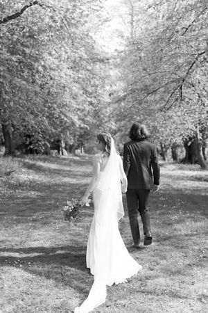 Black and white photo of bride and groom walking hand in hand through a tree-lined path on their wedding day.