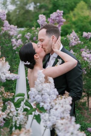 Bride and groom share a romantic kiss surrounded by blooming purple lilac bushes on their wedding day.