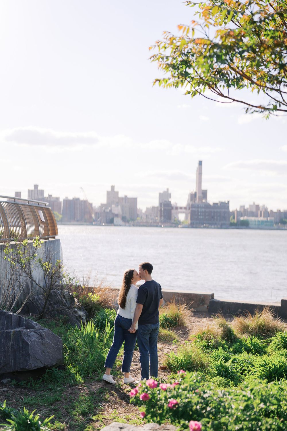 Couple kissing by the East River waterfront with lush greenery and NYC skyline in the background on a sunny day.