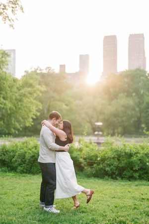 Couple embracing at golden hour in Central Park with lush greenery and NYC skyline in the background.