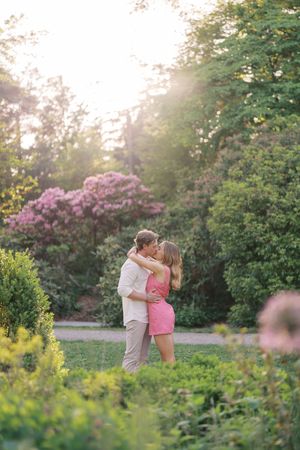 Couple sharing a romantic kiss in a lush green garden with pink blooming trees and golden sunset light.