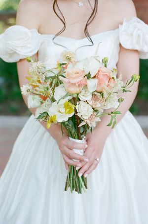 Bride in off-shoulder white gown holds a romantic bouquet of peach roses, white flowers, and greenery wrapped with ribbon.