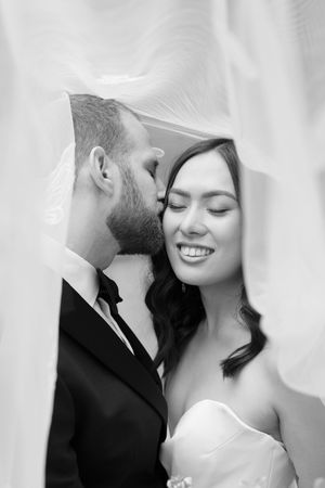 Black and white wedding photo of groom kissing bride on cheek under veil, bride smiling with eyes closed.