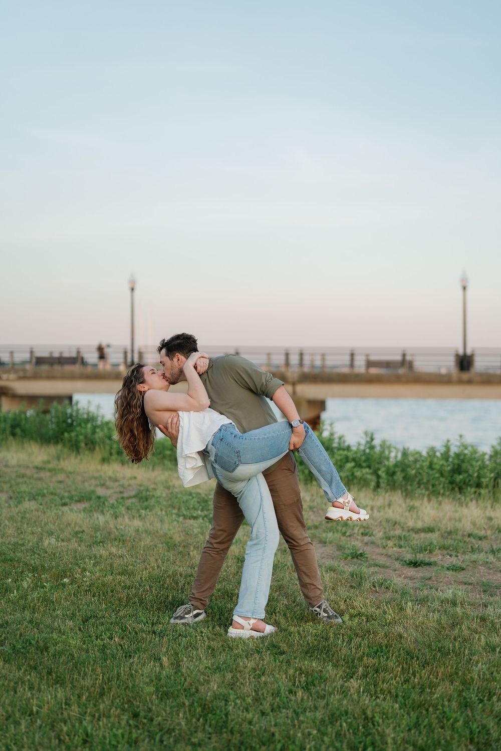 Couple sharing a romantic dip kiss on green grass near waterfront at sunset.