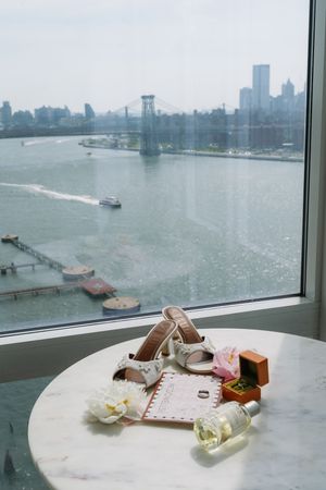 Bridal accessories including heels, rings, and perfume on marble table with NYC skyline and Williamsburg Bridge view.