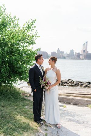 A bride and groom share a romantic moment by the waterfront with a lush green tree and city skyline backdrop.