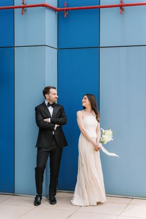A bride in a cream gown holding white flowers stands beside a groom in a black tuxedo against a blue wall.
