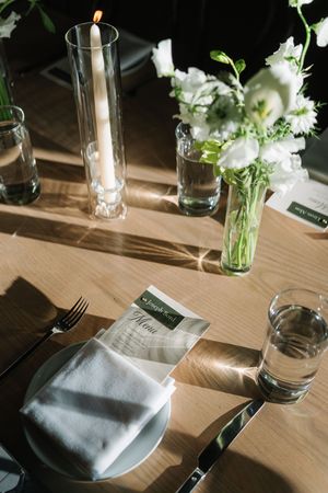 Elegant wedding table setting with white floral centerpiece, candle, folded napkin, menu card, and glassware on wooden table.