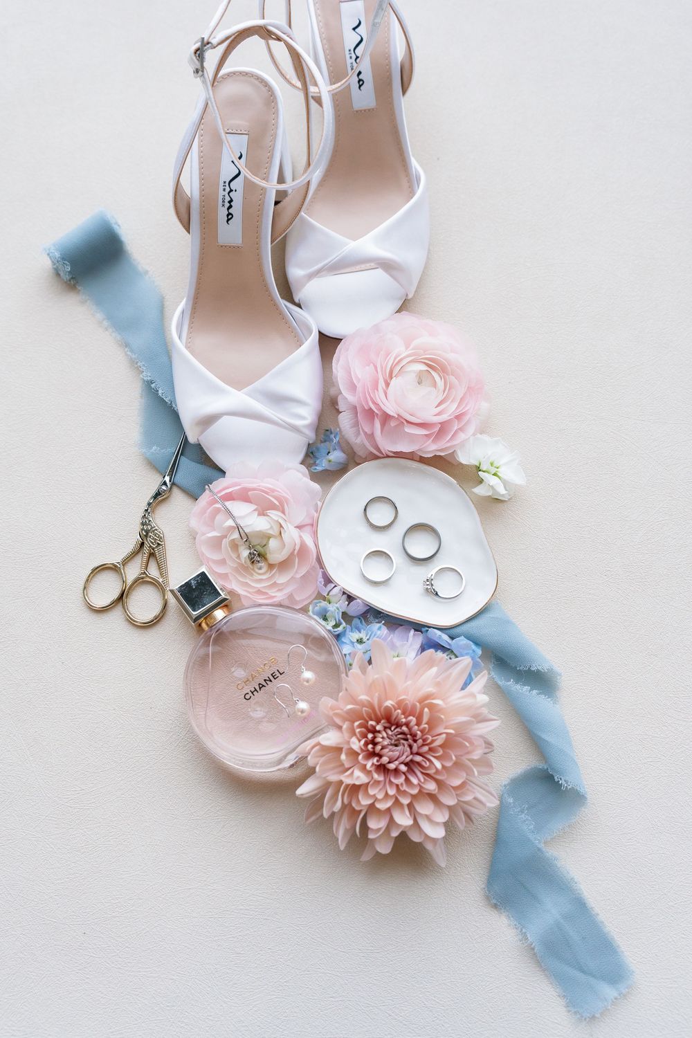 Elegant wedding flat lay with white heels, Chanel perfume, pink flowers, gold scissors, blue ribbon, and rings on a plate.