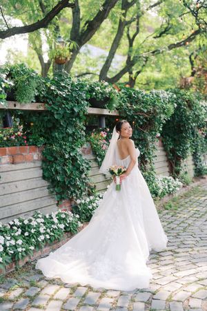 Bride in a white lace gown holding a bouquet, standing on cobblestone path beside an ivy-covered wooden fence.