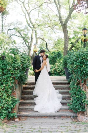 Bride and groom share a romantic embrace on ivy-covered stone steps in a lush garden setting.