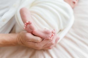 Adult hands gently cradling tiny newborn baby feet wrapped in a soft white blanket.