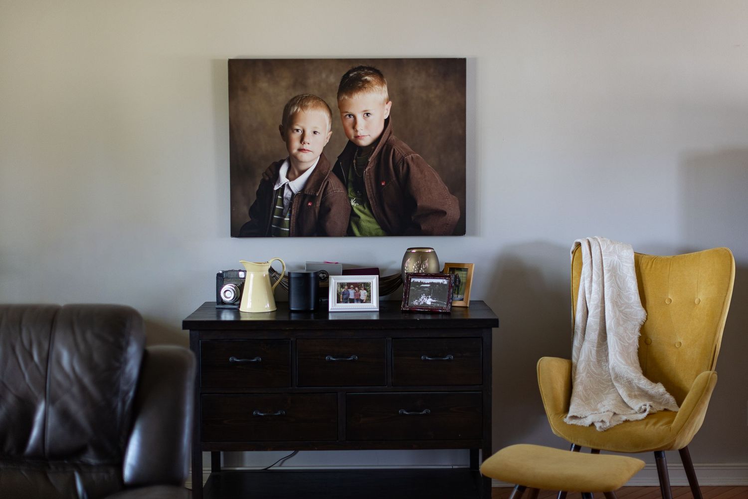 Framed portrait of two boys on wall above dark dresser with decor, beside yellow armchair with gray throw blanket.