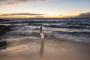 Woman in white dress walking into ocean waves at a beautiful beach sunset.