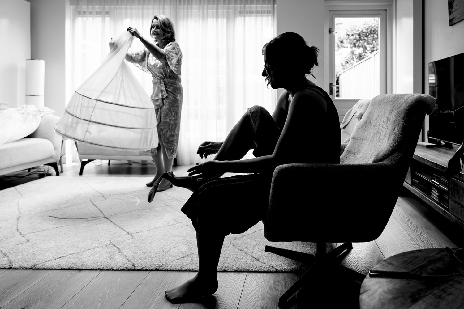 Intiem zwart-wit moment: bruid in silhouet bij het aankleden, moeder zichtbaar met de hoepelrok op de achtergrond