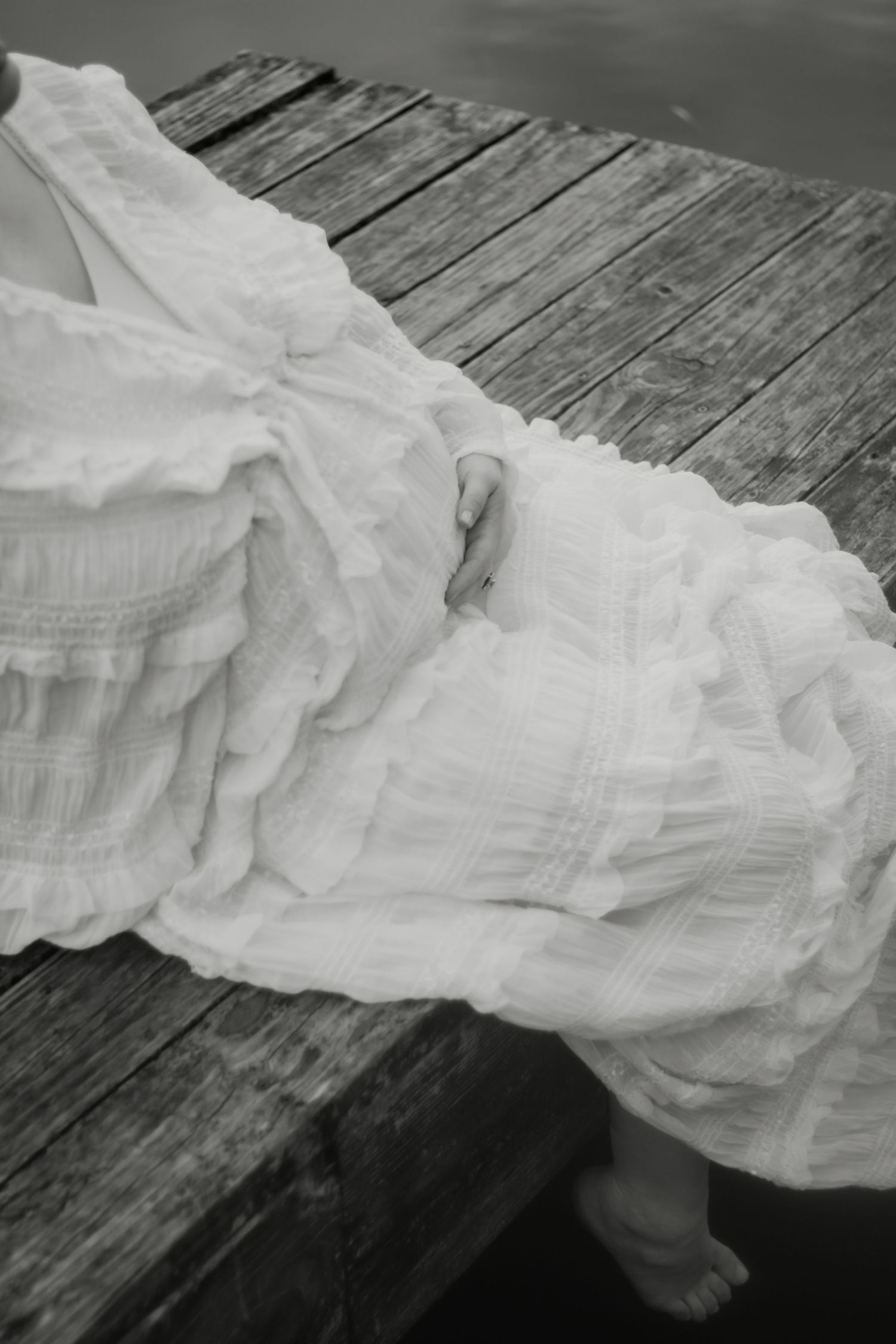 Black and white photo of a pregnant woman in a white ruffled dress sitting on a wooden dock.