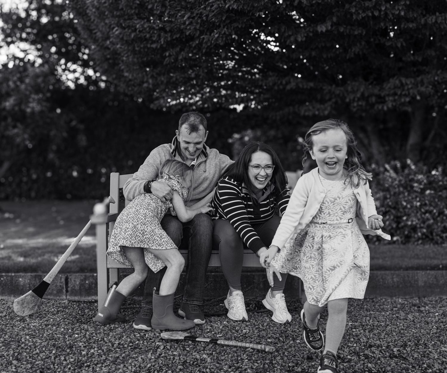 Black and white photo of a joyful family with two young girls laughing on a garden bench outdoors.