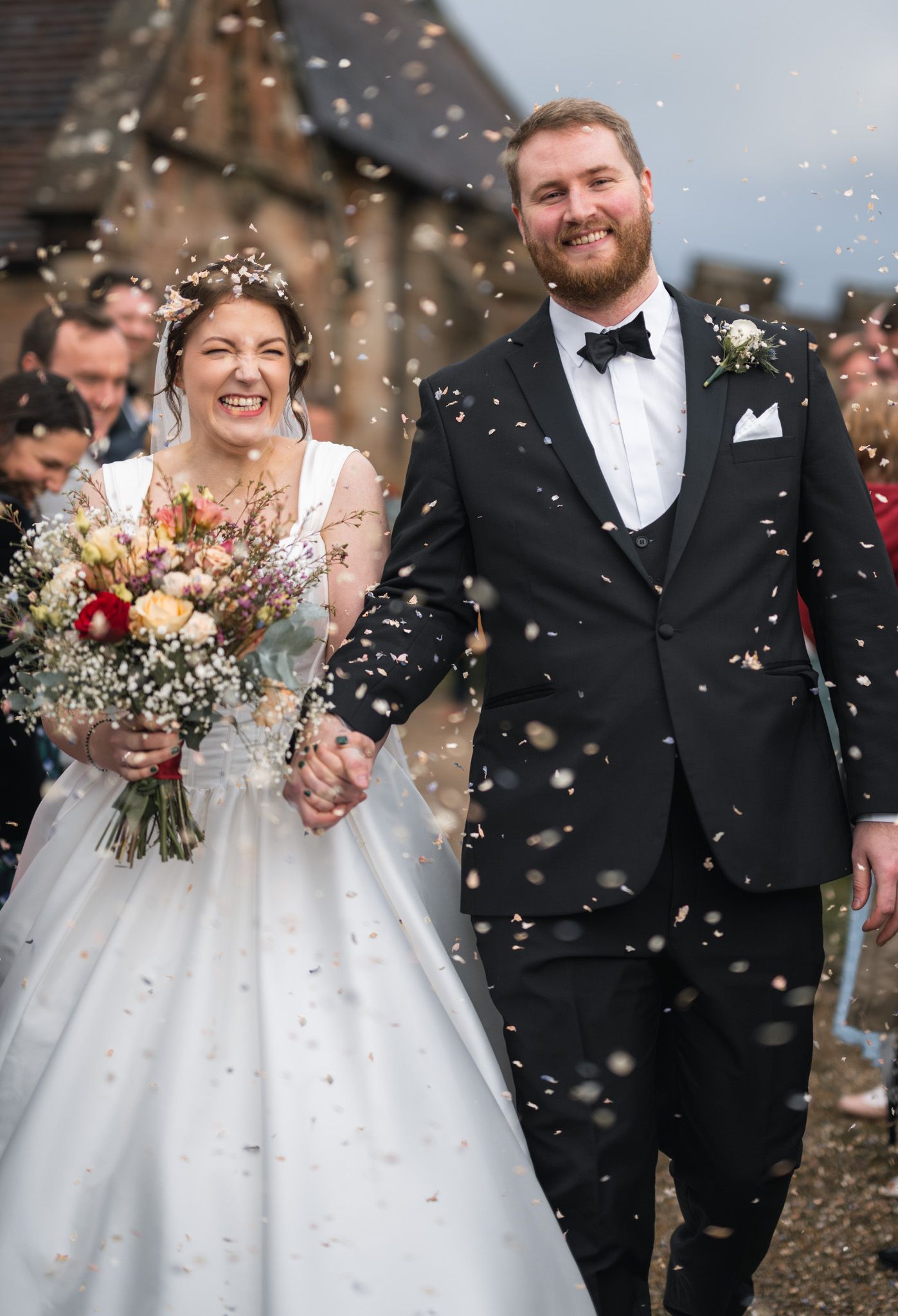 Laughing newlyweds celebrate with confetti outside a stone church, bride carrying a wildflower bouquet.