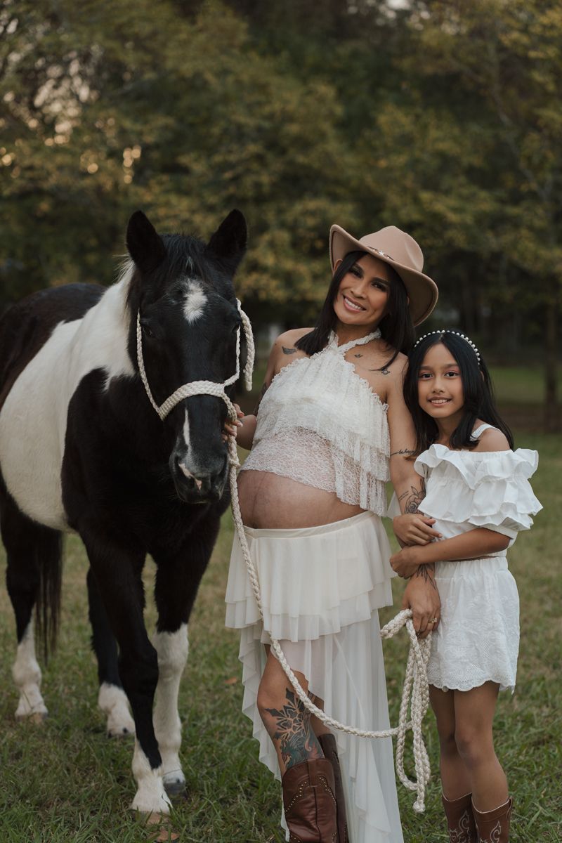 Pregnant woman in white dress and cowboy hat poses with daughter and black-and-white horse in outdoor field. maternity photoshoot Sydney