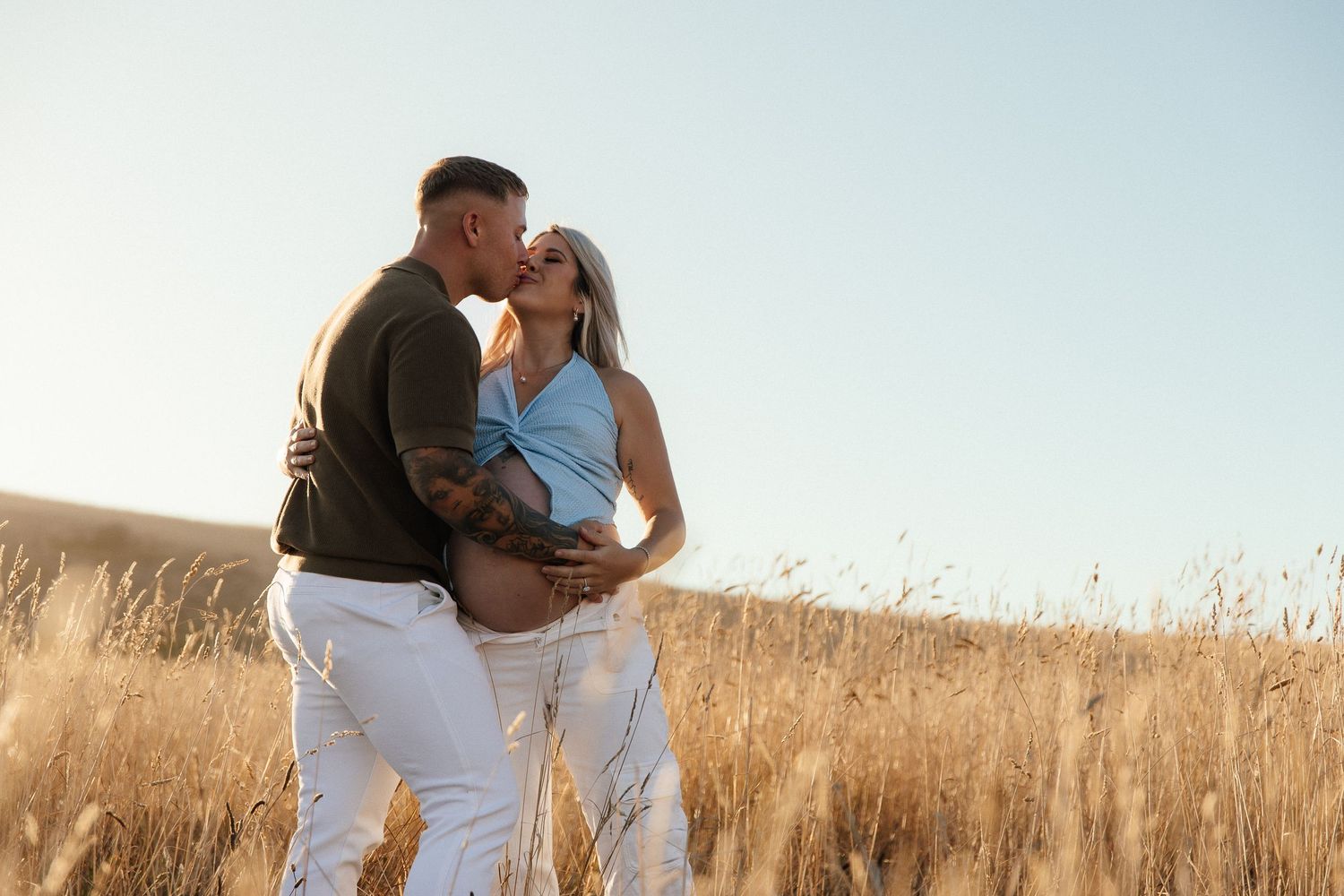Couple sharing a kiss in a golden wheat field, woman with baby bump wearing blue top and white pants on a sunny day.