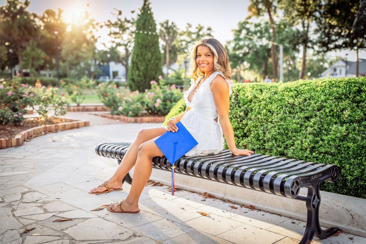 Young woman in white dress holding blue folder sits on ornate bench in sunny park garden.