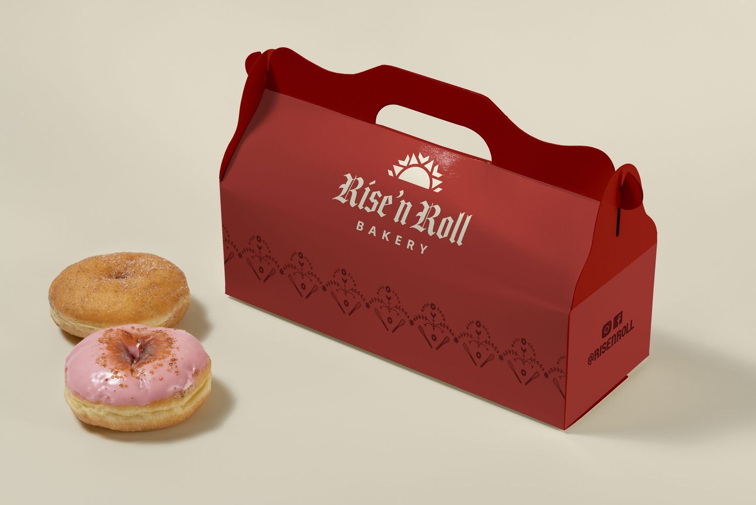 Red Rise n Roll Bakery gift box with handle displayed next to two frosted donuts on a neutral background.
