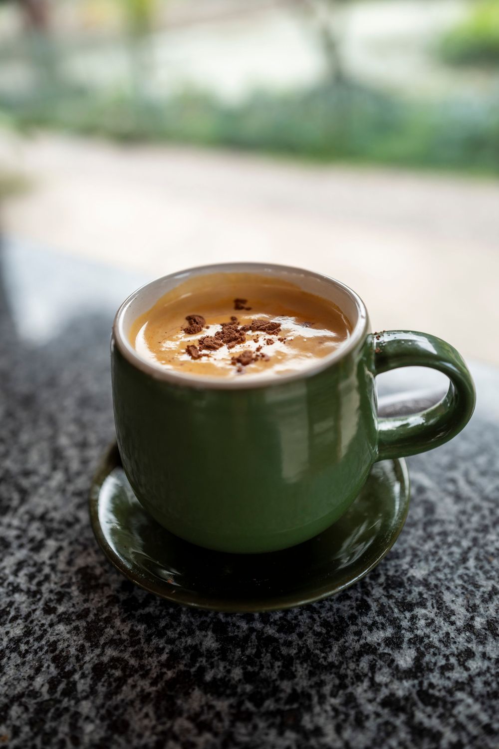 A green ceramic coffee mug with frothy cappuccino topped with chocolate shavings, resting on a matching saucer.