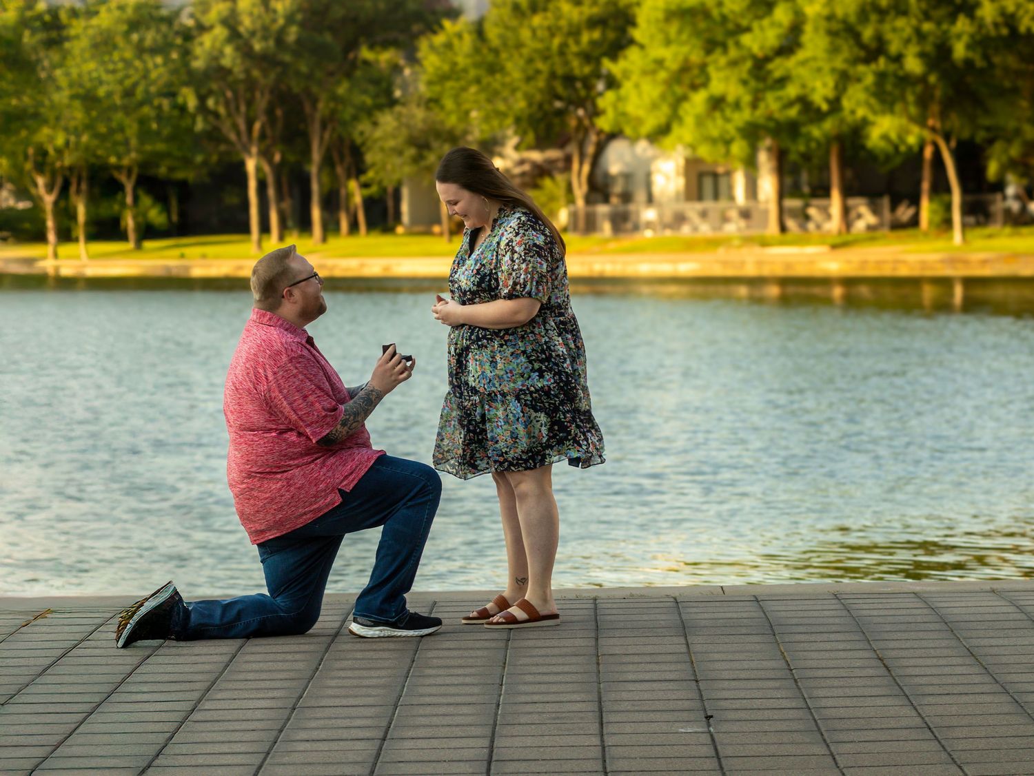 Man proposes on one knee to woman on a wooden dock by a serene lake surrounded by lush green trees.
