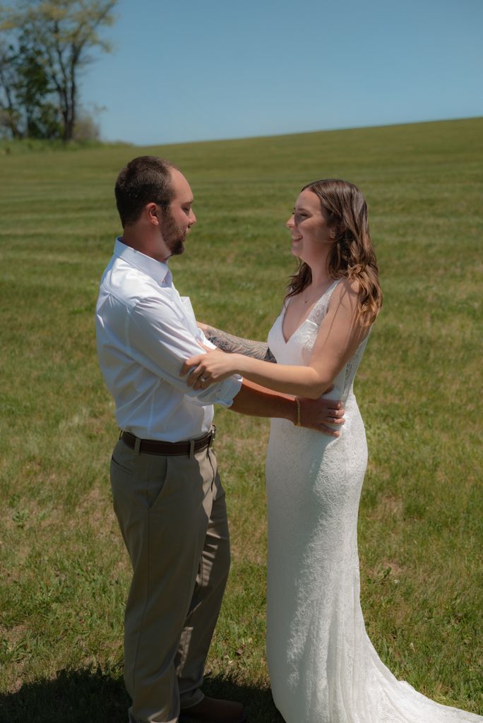 A bride in a white gown and groom in tan pants share a tender first look moment on a sunny green hillside.