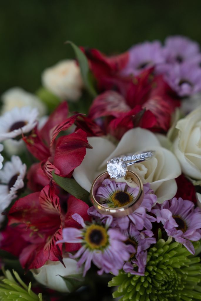 Wedding rings resting on a colorful bridal bouquet with red alstroemeria, white roses, and purple daisies.