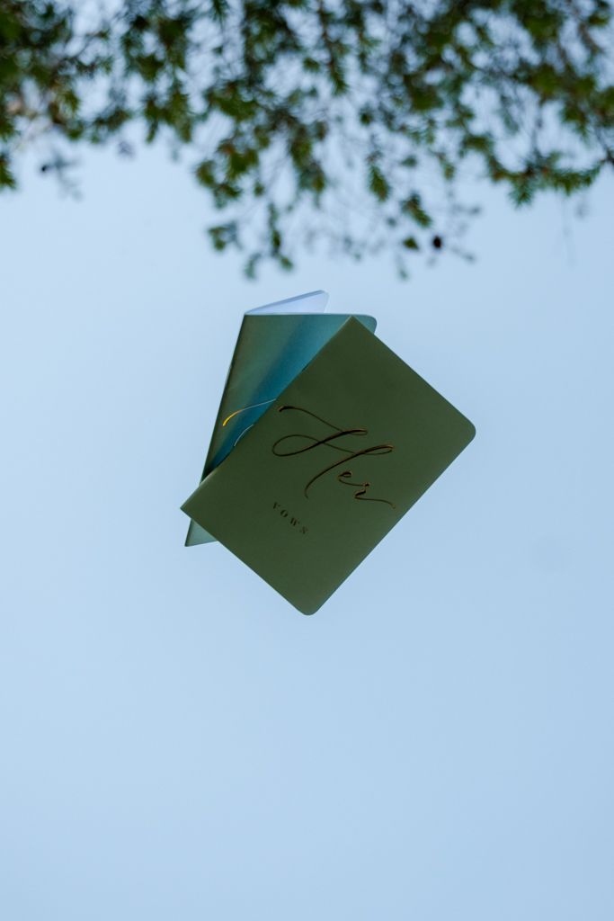 Two green wedding vow books with elegant script float against a clear blue sky beneath tree branches.