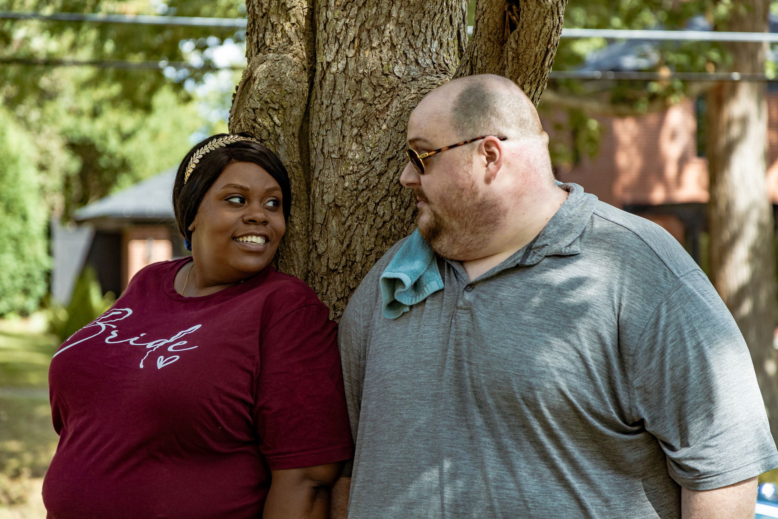 A couple smiles at each other outdoors near a tree, woman wearing a burgundy bride shirt, man in gray polo.