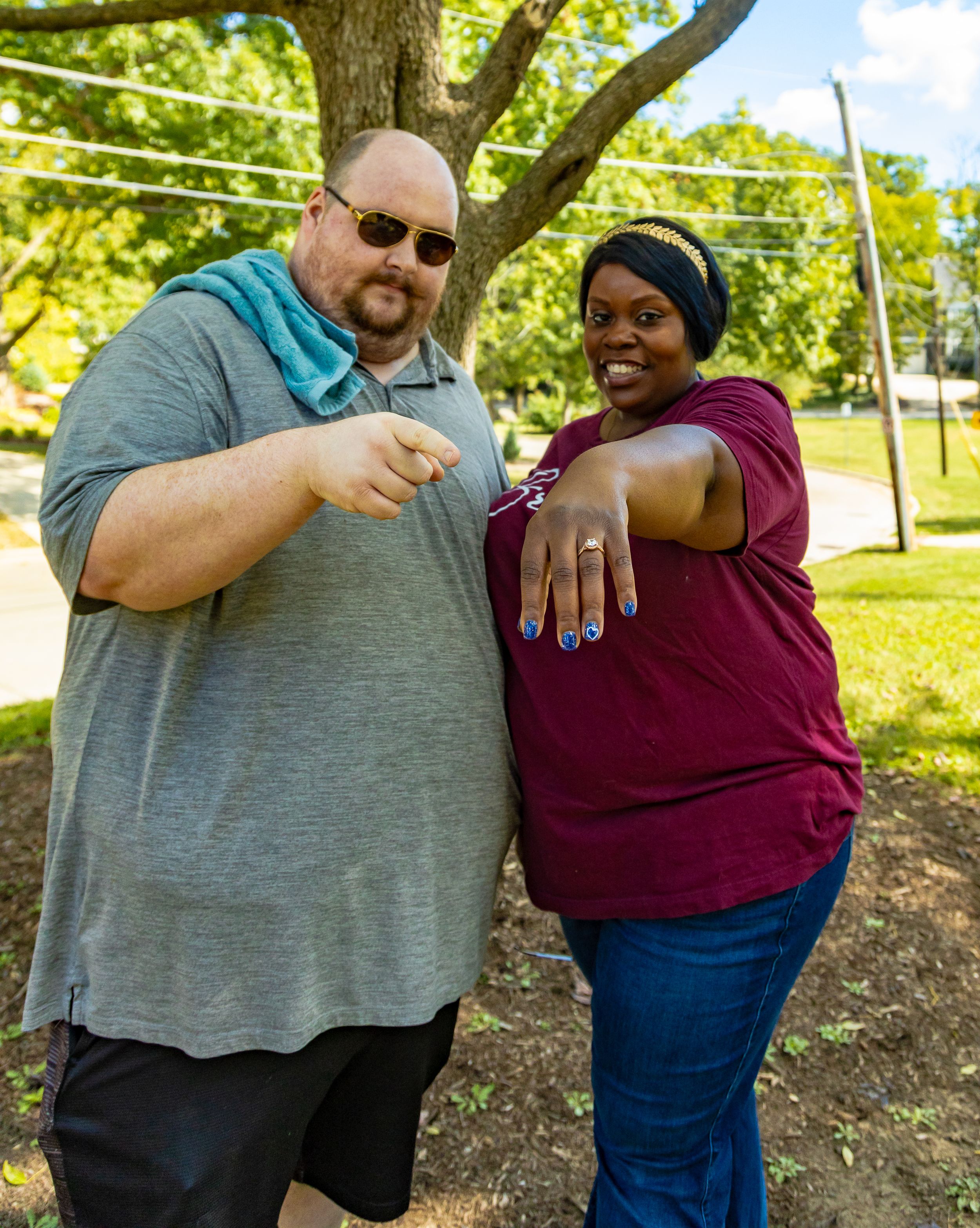 Couple smiling outdoors showing off engagement ring, woman in maroon top with blue nails, man in gray shirt, trees in background.