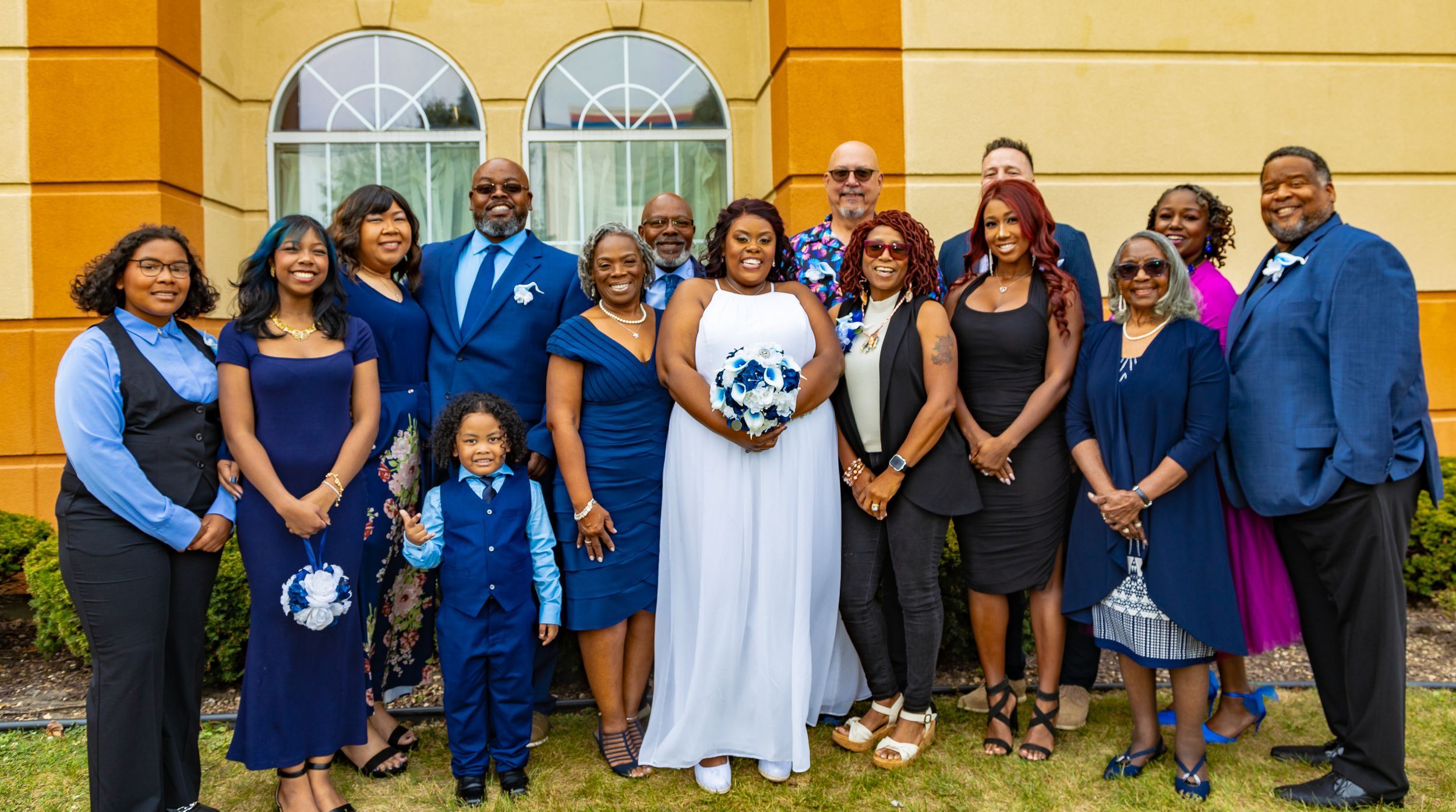 Wedding group photo with bride in white gown, groom, and family members in blue attire outside yellow building.