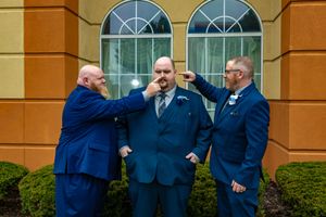 Three groomsmen in blue suits joke around outside a yellow building before a wedding ceremony.