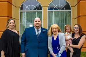 Family group of five posing formally outdoors, including a man in blue suit, women in elegant attire, and a child being held.