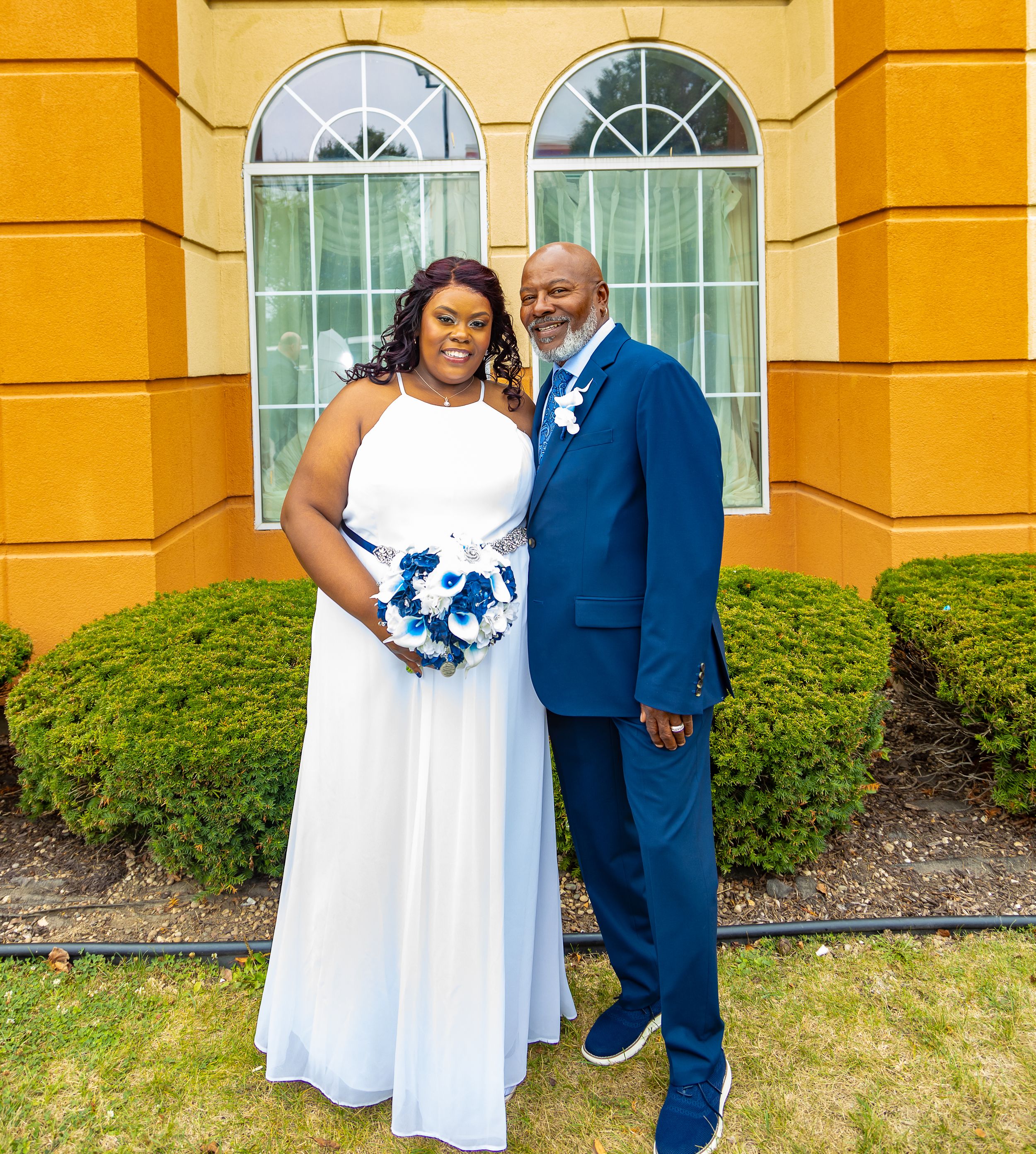 Bride in white gown and groom in blue suit pose together outside yellow building on their wedding day.