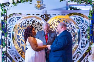 Interracial couple exchanging vows at wedding altar with ornate white heart backdrop and officiant.